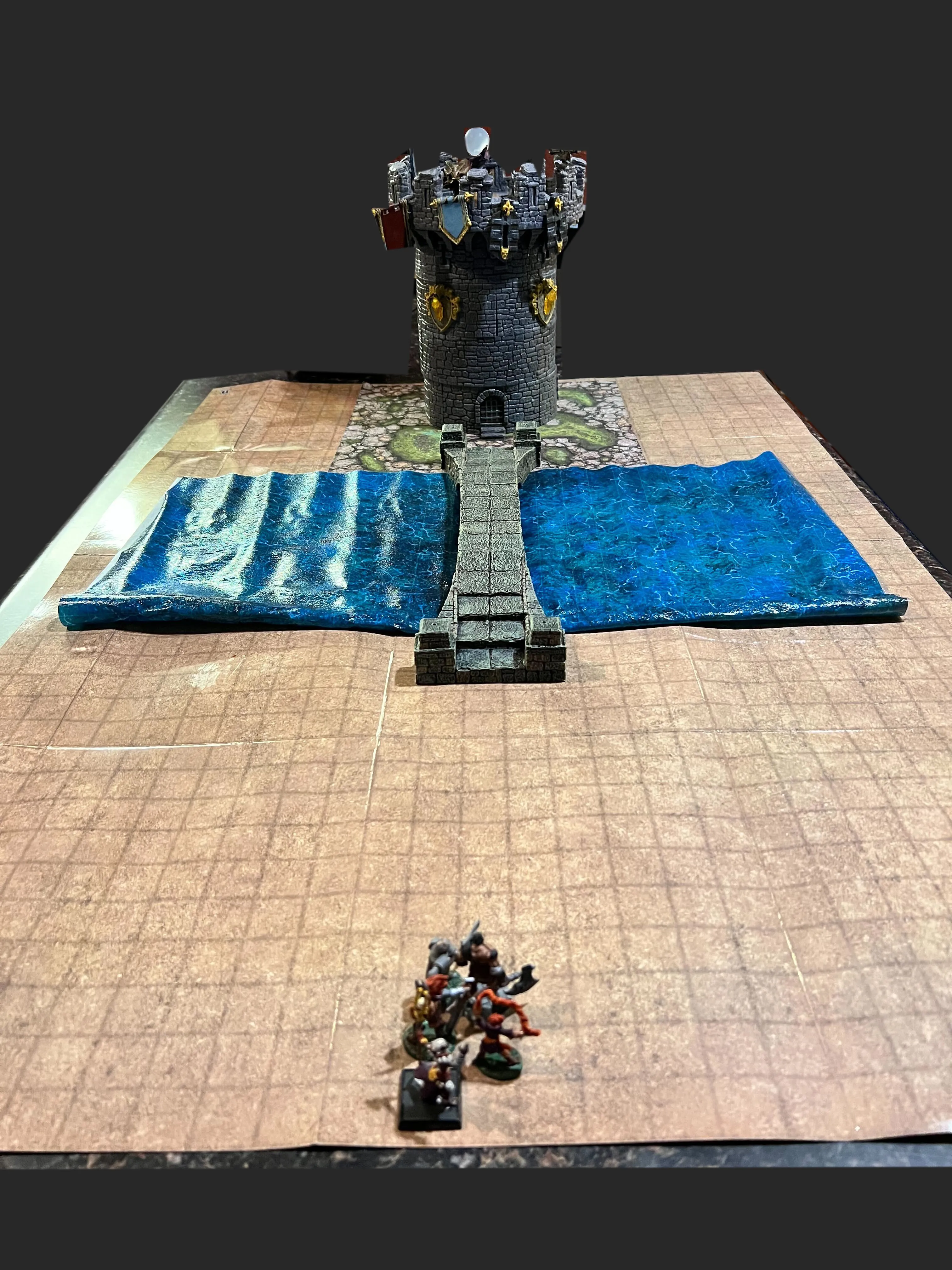 The party approaches the tower! A group of miniatures approaches a watchtower on a gridded map with a body of water and a bridge.